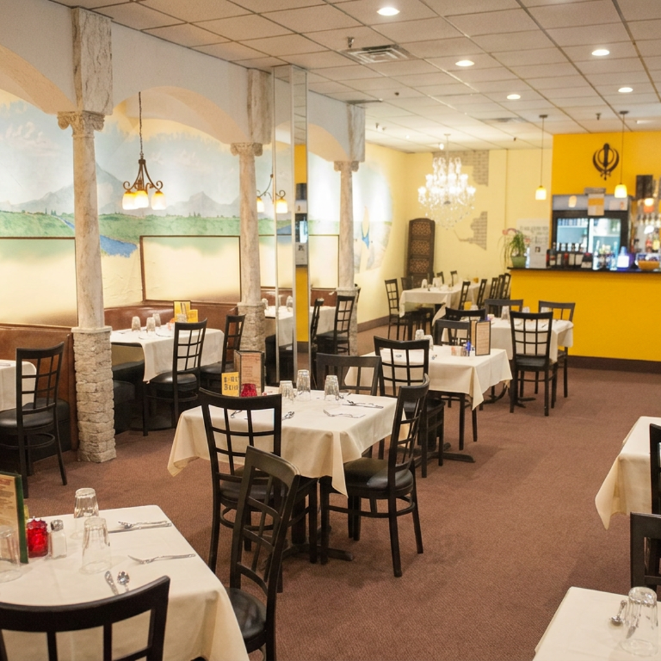 Bright Indian restaurant dining room with white tablecloths, black chairs, mural, decorative columns, and yellow Tandoor bar.