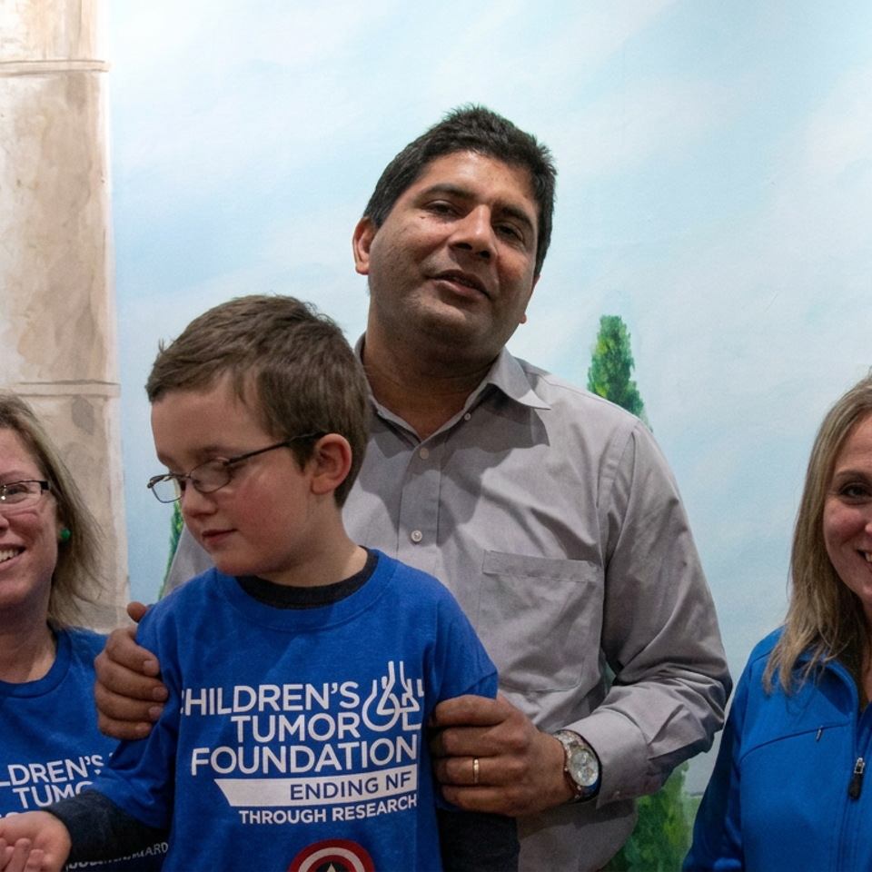 Two women, a boy, and a man pose together indoors wearing blue Children’s Tumor Foundation shirts before a painted outdoor backdrop.