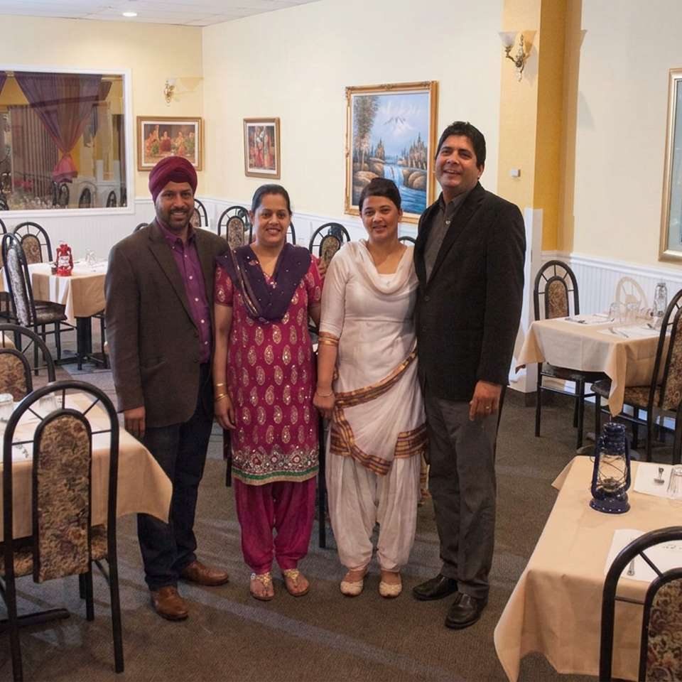 Four people pose inside a warmly decorated restaurant dining room with set tables, patterned chairs, and framed artwork.