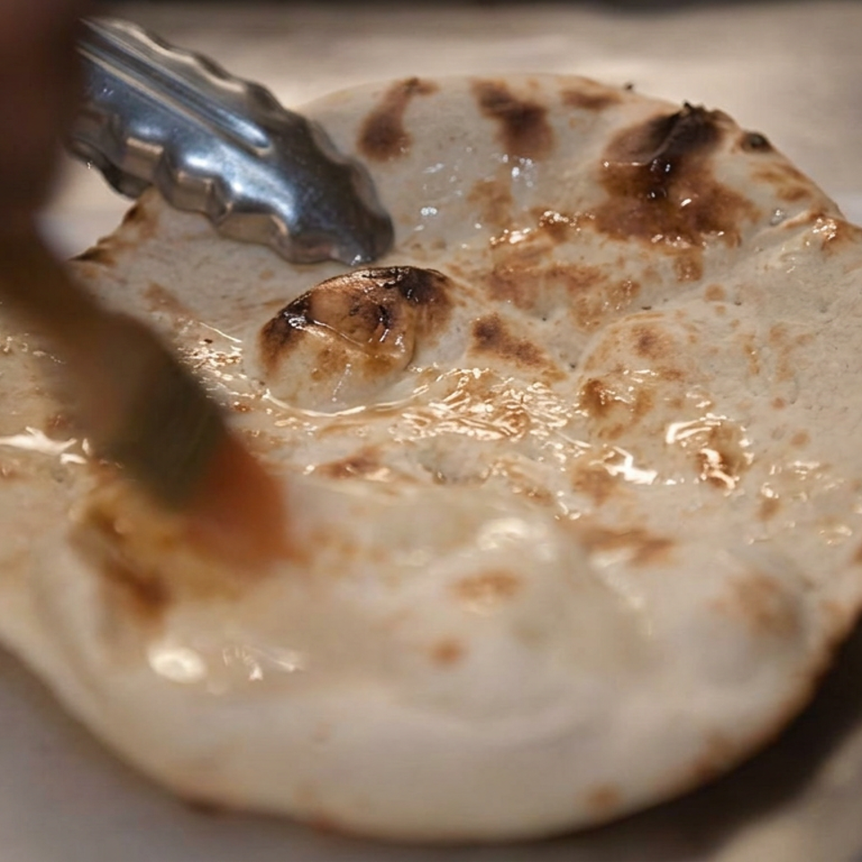 Grilled flatbread with browned spots glistens with butter as metal tongs lift its edge on a paper-lined surface.